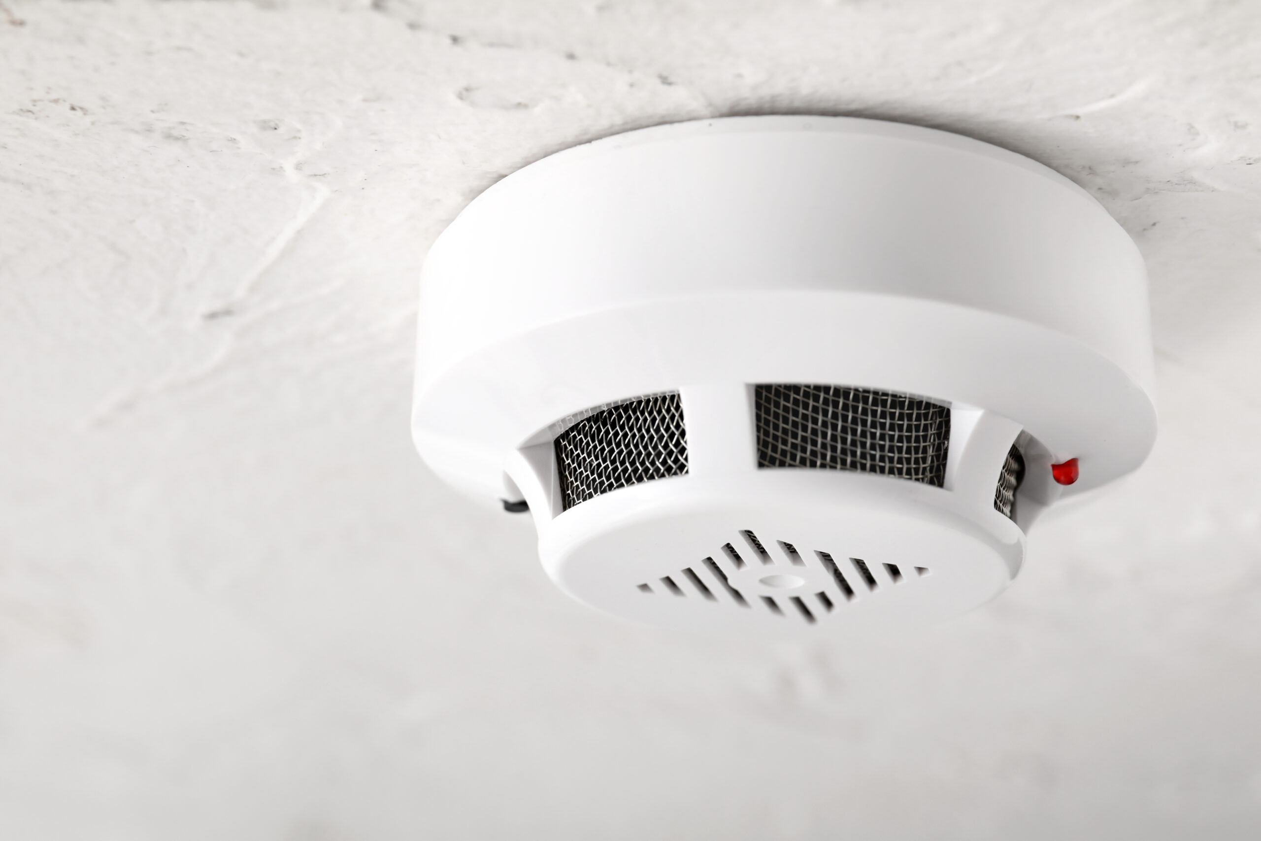 A white smoke detector, crucial in carbon monoxide research, is mounted on a textured ceiling. The device features a circular design with air vents and a small red indicator light on the side.