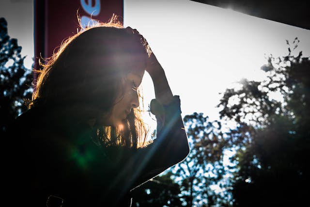 A silhouette of a person touching their head stands against a bright background with sunlight streaming through trees. The light creates a lens flare effect, adding an ethereal feel to this moment, subtly reflecting the inner contemplation often felt in the shadow of intimate partner violence.