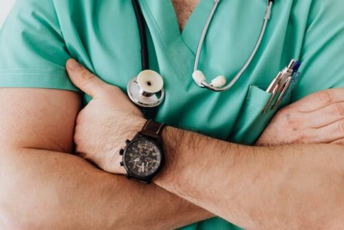 A person in green scrubs stands with arms crossed, ready to offer advice. A stethoscope hangs around their neck, and a pen peeks from the shirt pocket. The individual, marked by their trusty wristwatch on the left wrist, exudes an air of calm amidst any accident's chaos.